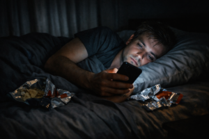 A person scrolling on their phone late at night in a darkened bedroom, representing the overstimulation cycle that makes everything feel boring.