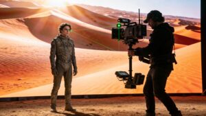 Cinematographer aiming a digital cinema camera at an actor standing in front of a giant curved LED volume displaying a realistic landscape.