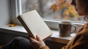 Person reading a paperback by a rainy window with a mug of tea.